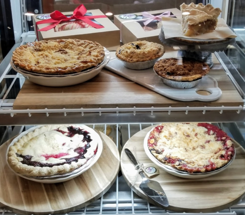 A display of various pies, including fruit and cream varieties, on wooden boards in a glass case.