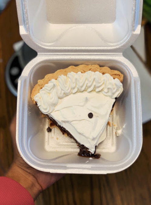 A slice of chocolate pie with whipped cream in a takeout container, held by a hand.