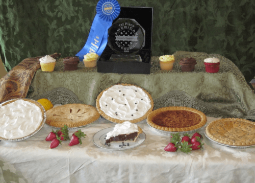 A display of various pies and cupcakes, featuring a blue ribbon and a trophy for first place in a baking competition.