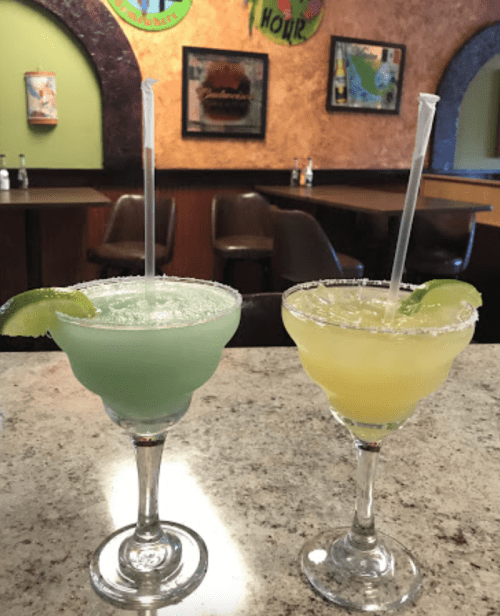Two colorful margaritas on a granite countertop, one green and one yellow, garnished with lime slices.