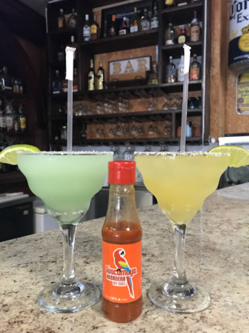 Two colorful margaritas in glasses with lime slices, accompanied by a bottle of hot sauce on a bar counter.