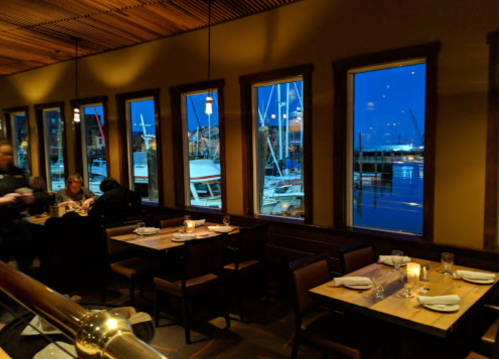 Cozy restaurant interior with wooden tables, dim lighting, and large windows showcasing a harbor at dusk.