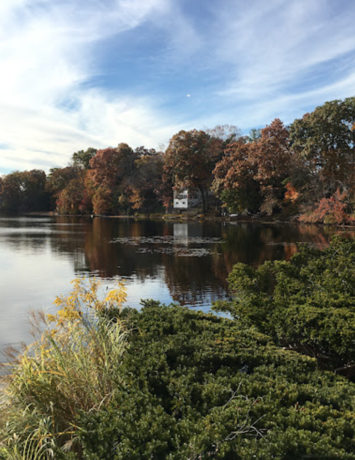 A serene lake scene with autumn foliage, a small white house, and lush greenery in the foreground under a blue sky.