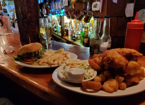 A burger with fries and a plate of fried shrimp with coleslaw, served at a bar with a variety of drinks in the background.