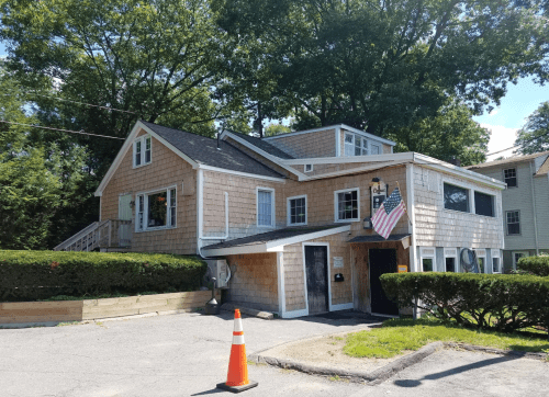 A two-story house with wooden shingles, an American flag, and a landscaped yard, surrounded by trees.