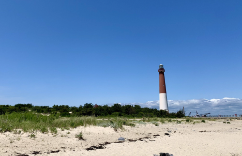 A tall lighthouse stands on a sandy beach under a clear blue sky, surrounded by green vegetation.