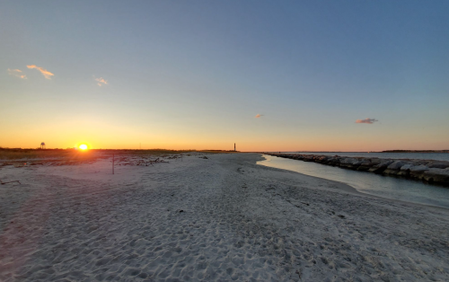 Sunset over a sandy beach with a calm waterway and distant lighthouse, creating a serene coastal scene.