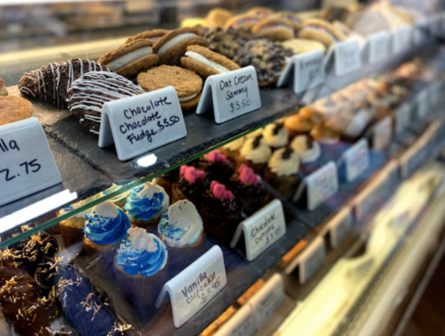 A display case filled with various cookies and cupcakes, each labeled with names and prices.
