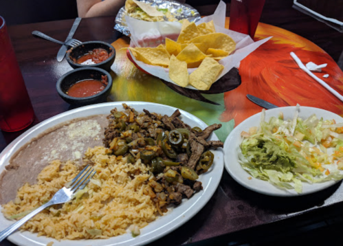 A plate of beef fajitas with rice and beans, alongside a salad and tortilla chips with salsa on a colorful table.