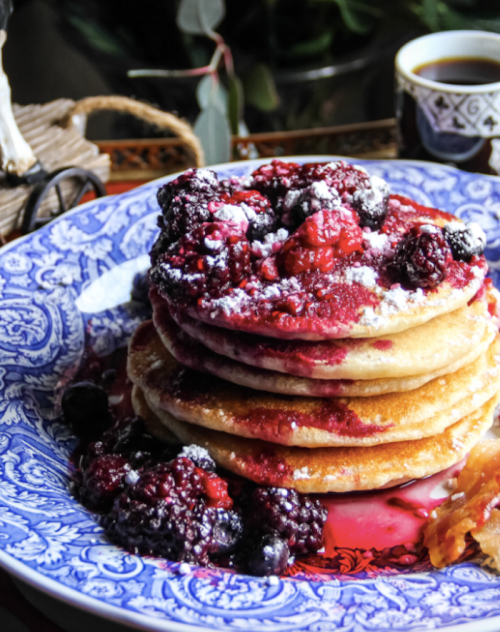 A stack of fluffy pancakes topped with mixed berries and powdered sugar, served on a decorative blue plate.