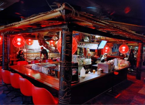 A vibrant bar with bamboo decor, red lanterns, and a lively atmosphere, featuring patrons and a well-stocked counter.