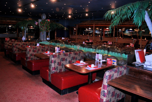 Interior of a restaurant featuring red booths, wooden tables, and tropical decor with palm trees and dim lighting.