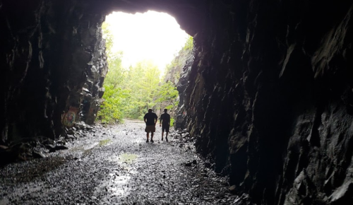 Two silhouetted figures walk through a rocky tunnel, with greenery visible at the tunnel's opening.