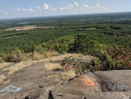 A panoramic view from a rocky outcrop overlooking a lush green landscape and distant water bodies under a blue sky.