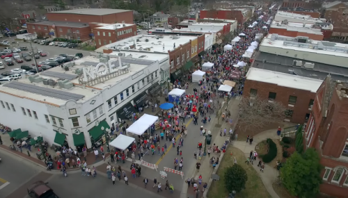 Aerial view of a bustling street festival with crowds, tents, and shops lining the street.