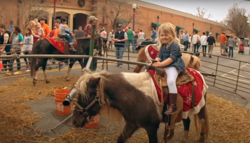 A young girl smiles while riding a pony at a festive outdoor event with people and decorations in the background.
