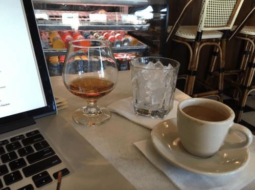 A laptop on a table beside a glass of whiskey, a glass of ice water, and a small cup of coffee.