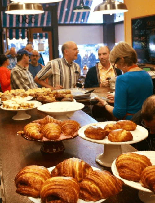 A busy café scene with people chatting at the counter and a display of pastries, including croissants and muffins.