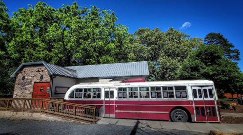 A vintage red and white bus parked beside a stone building with a gray roof, surrounded by green trees.
