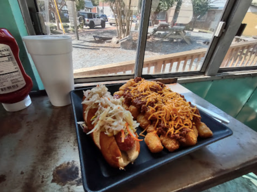 A plate with two hot dogs topped with coleslaw and chili, alongside a cup, with a view of an outdoor area in the background.