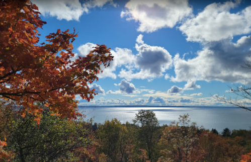 A scenic view of a lake surrounded by colorful autumn trees under a bright blue sky with fluffy clouds.