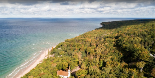 Aerial view of a coastline with sandy beach, lush green trees, and calm blue waters under a cloudy sky.