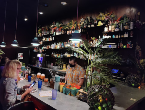 A vibrant bar scene with tropical decor, a bartender preparing drinks, and a customer using a phone at the counter.