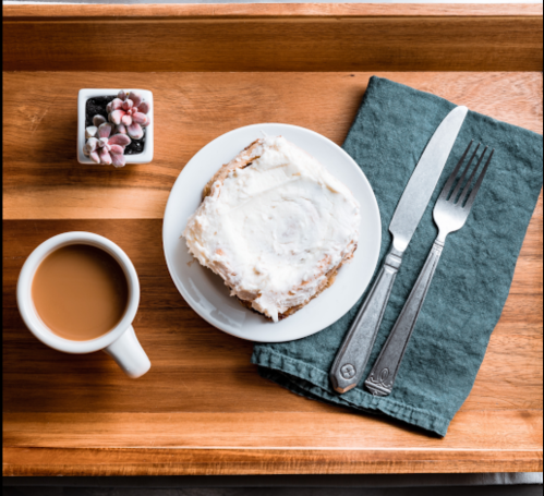 A wooden tray holds a cup of coffee, a slice of frosted cake, and a fork and knife on a green napkin.