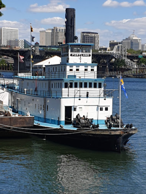 A historic tugboat docked on a calm waterway, with a city skyline in the background under a clear blue sky.
