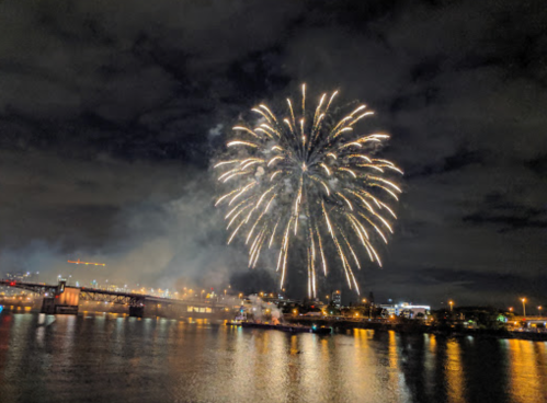 Colorful fireworks burst over a city skyline at night, reflecting on the water below.