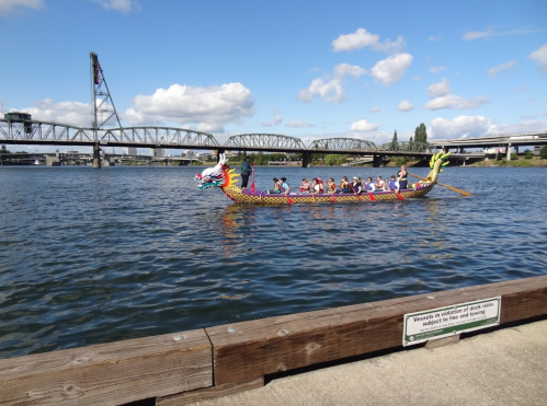 A dragon boat with paddlers glides across a river under a bridge, with blue skies and clouds above.