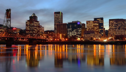 City skyline at dusk, with illuminated buildings reflecting on a river, and a bridge in the foreground.
