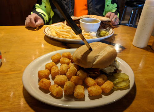 A plate with a sandwich, tater tots, and pickles, with another plate of fries and a drink in the background.