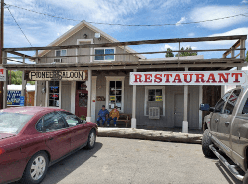 A rustic building with signs for "Pioneer Saloon" and "Restaurant," featuring two people sitting outside.