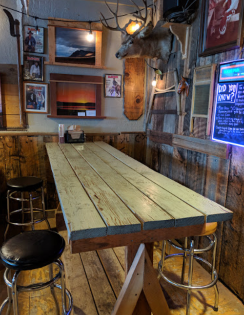 A rustic bar area with a wooden table, stools, and mounted deer head on the wall, surrounded by framed pictures.