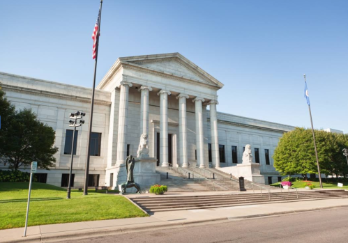A large, classical-style building with columns, flanked by statues, surrounded by green grass and trees.