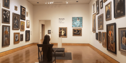 A gallery room featuring various framed portraits on the walls, with a viewer seated in front of the artwork.