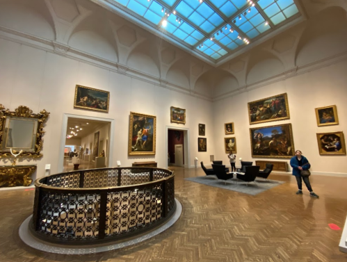 A spacious art gallery featuring large paintings, elegant decor, and a visitor standing near a seating area.
