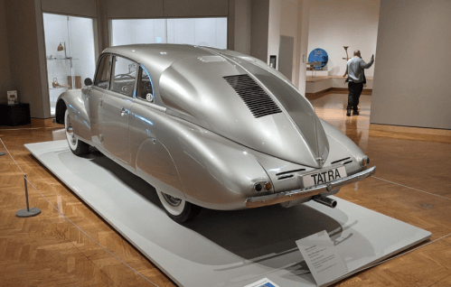 A vintage silver Tatra car displayed in a museum, with a person in the background admiring the exhibits.