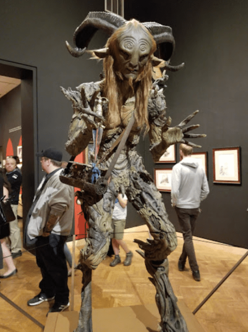 A tall, intricate sculpture of a creature with horns and long hair, surrounded by people in an art gallery.