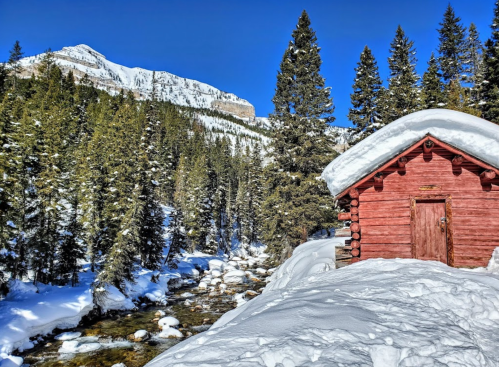 A red cabin covered in snow beside a stream, surrounded by evergreen trees and mountains under a clear blue sky.