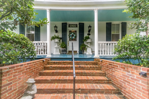 A charming porch with blue ceiling, white columns, and brick steps, adorned with plants and a festive wreath.