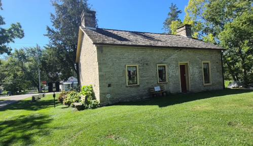 A small stone house with a sloped roof, surrounded by green grass and trees, set in a peaceful outdoor environment.