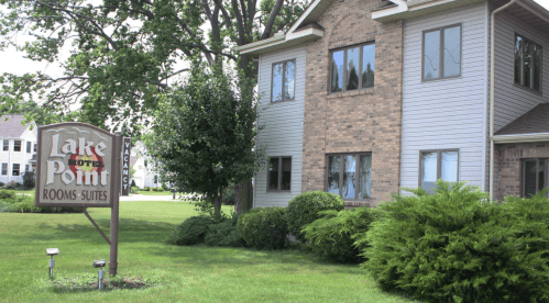 A brick building with large windows and a sign reading "Lake Point Rooms-Suites" surrounded by green grass and trees.