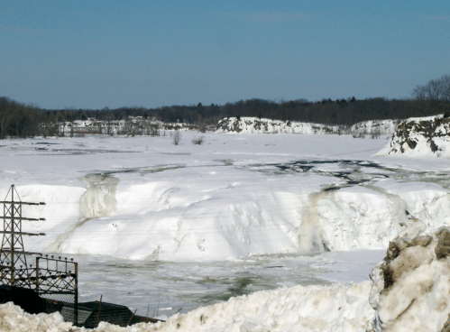 A snowy landscape with a frozen river and distant trees under a clear blue sky. Power lines are visible in the foreground.