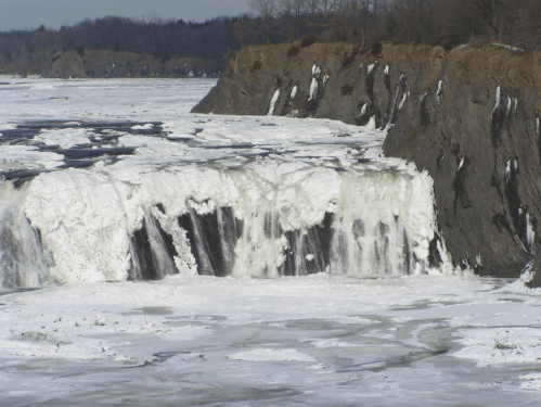A frozen waterfall cascading over rocky cliffs, surrounded by icy terrain and a winter landscape.