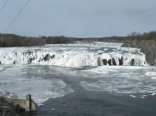 Frozen waterfall cascading over rocky cliffs, surrounded by icy river and barren trees under a cloudy sky.