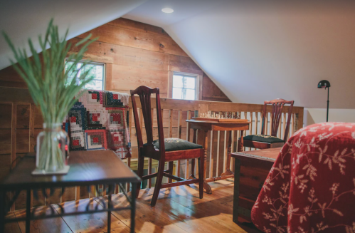 Cozy attic space with wooden walls, a chess set, chairs, and a table, featuring a red blanket and a vase of greenery.