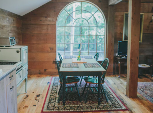 Cozy kitchen with a wooden dining table, chairs, and large arched window, surrounded by rustic decor and warm lighting.