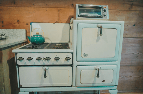 A vintage white stove with a teal kettle on top and a small toaster oven beside it, set against a wooden wall.